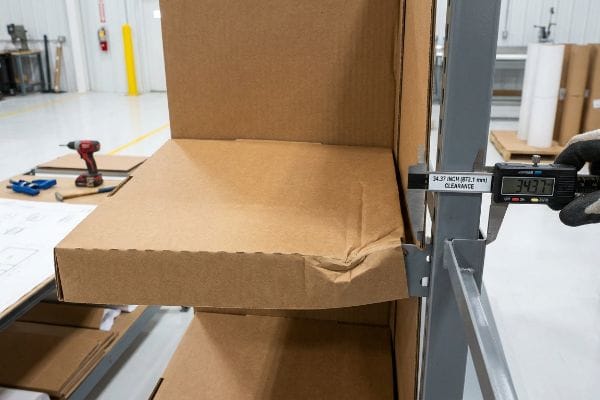 Medição da folga da gôndola Um paquímetro digital mede 873,1 mm (34,37 polegadas) de folga em uma prateleira de aço de gôndola, mostrando uma fixação de papelão na extremidade.