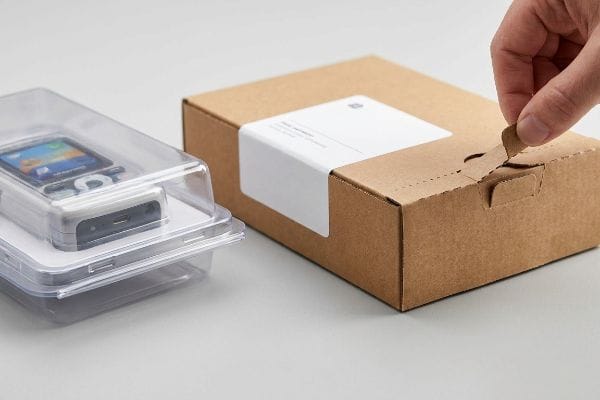Brown corrugated frustration-free packaging box with a tear strip being opened, next to a clear plastic clamshell holding an electronic device.