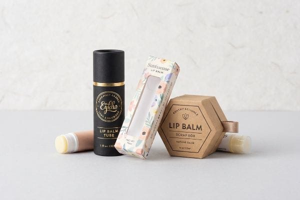 Custom Lip Balm Packaging Three custom lip balm packaging designs: black Egkho Lip Balm Tube with gold foil, floral Sundisouse Lip Balm window box, and hexagonal Beachy Science Lip Balm Scrap Box.