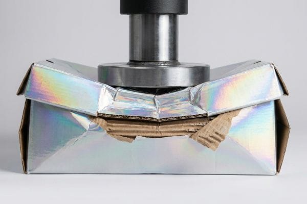 Holographic foil-laminated corrugated box imploding under hydraulic BCT pressure, revealing crushed flutes.