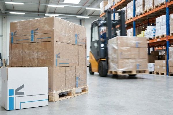 Warehouse Forklift Moving Boxes A yellow forklift transports shrink-wrapped boxes on a pallet in a busy warehouse, with stacks of 'IK' branded cardboard packaging in the foreground.