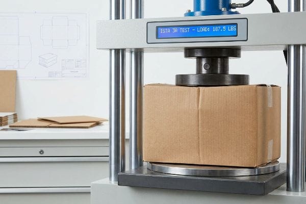 Brown cardboard box undergoing an Ista 3a Test on a hydraulic press, displaying a load of 187.5 Lbs. Packaging blueprints visible.