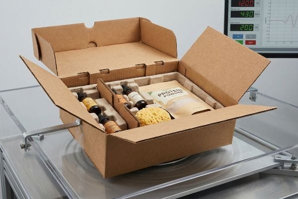Brown corrugated box with glass bottles, Protein Powder, and sponge, undergoing ISTA 3A vibration testing.