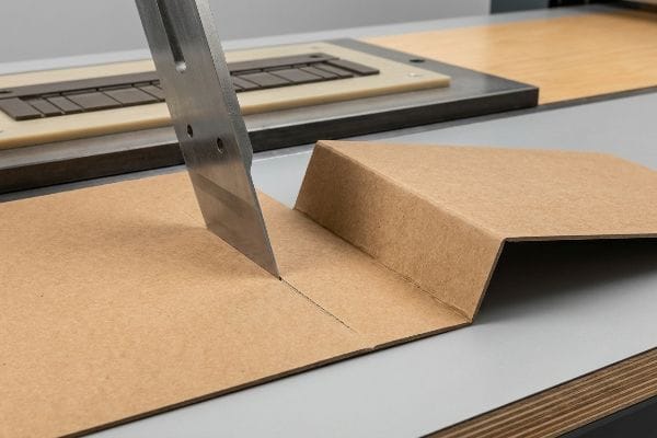 Brown virgin kraft linerboard receives a precise score from a steel rule die, forming a clean fold line.