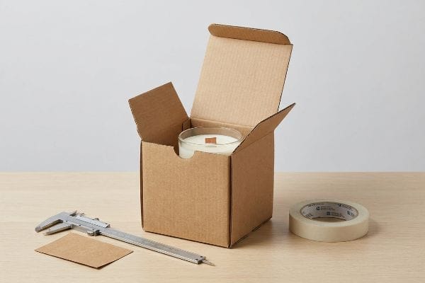 Candle Box Measurement Tools Corrugated kraft box with a white glass candle, a silver caliper, and masking tape on a light wood table, for precise packaging measurements.