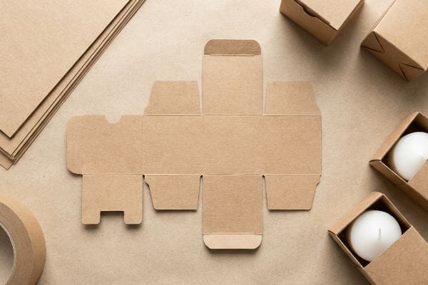 Kraft Candle Box Template An overhead view displays an unfolded kraft paper box template, kraft tape, and two small kraft boxes with white candles, emphasizing eco-friendly packaging.