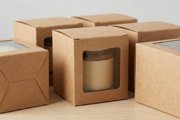 Natural brown kraft paper boxes with clear die-cut windows, one revealing a glass jar with a blank label, ideal for sustainable packaging.