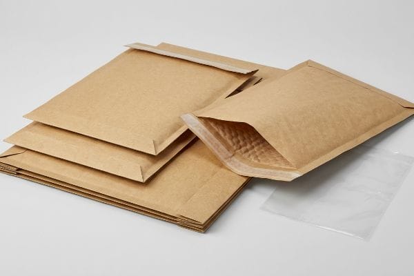 Kraftpapier-Versandtaschen (gepolstert) Gepolsterte Versandtaschen aus braunem Kraftpapier, eine davon geöffnet, sodass die Luftpolsterfolie sichtbar ist, neben einem durchsichtigen Polybeutel, die umweltfreundliche Verpackungsoptionen aufzeigen.
