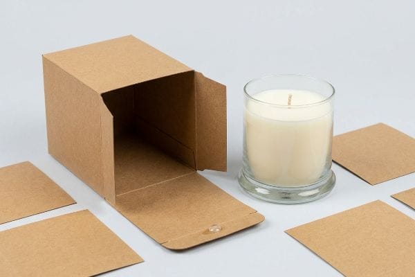Kraft Candle Box Packaging Unlined kraft paperboard candle box, open, with a clear closure dot, next to a white candle in a glass jar and flat kraft sheets.