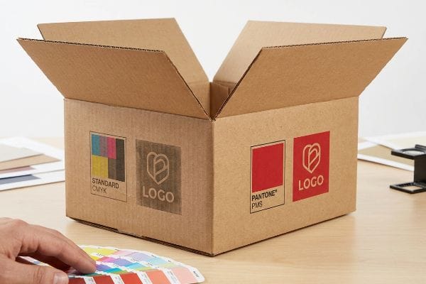 CMYK vs PMS Printing Corrugated Kraft box demonstrating Standard CMYK's faded logo versus Pantone PMS's vibrant logo, with a hand holding a color swatch book.