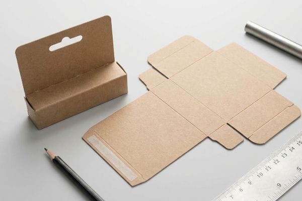 Kraft Packaging Dieline Packaging design process with a kraft paper lip balm box, hang tab, and its unfolded dieline on a grey desk.
