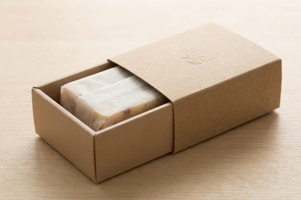 Two artisanal soap bars, wrapped, partially sliding from a Kraft paperboard drawer box with an embossed logo.
