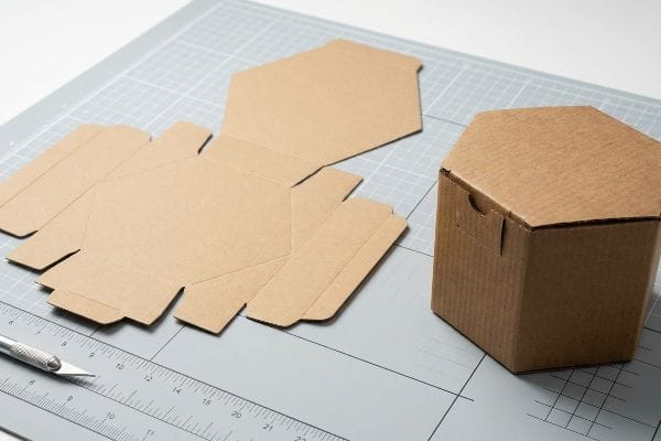 Hexagon Box Assembly Dieline An assembled brown kraft paper hexagon box stands next to its flat dieline on a grey cutting mat with a craft knife.