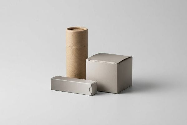 Eine minimalistische Darstellung von drei leeren Kosmetikverpackungsmodellen: eine braune Kraftpapiertube, eine hellgraue Schiebebox und eine quadratische Schachtel.