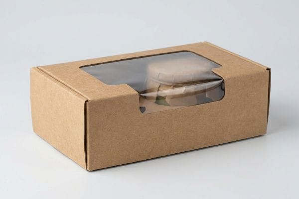 A brown kraft retail packaging box features a clear die-cut window, showcasing two small, round containers inside.