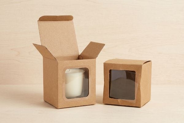 Brown kraft paper window cartons, one open with a white candle, one closed, featuring die-cut viewing windows.