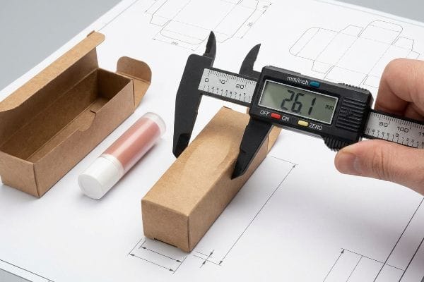 Caliper Lip Balm Box Measurement A digital caliper precisely measures a kraft paper lip balm box to 26.1 mm, alongside a cosmetic tube and packaging blueprint.