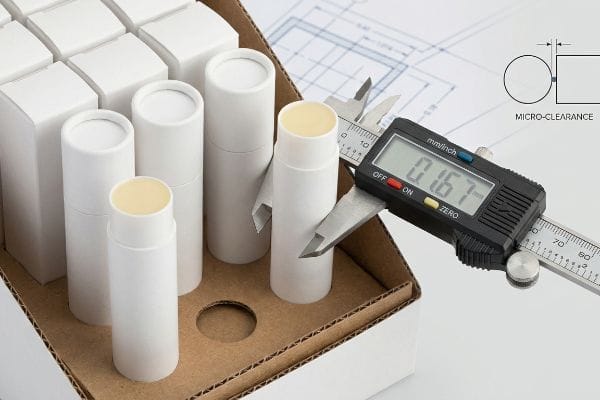 Lip Balm Micro-Clearance A digital caliper measures white cylindrical lip balm tubes, showing 0.167 mm/inch, highlighting micro-clearance in a corrugated tray with a technical blueprint.