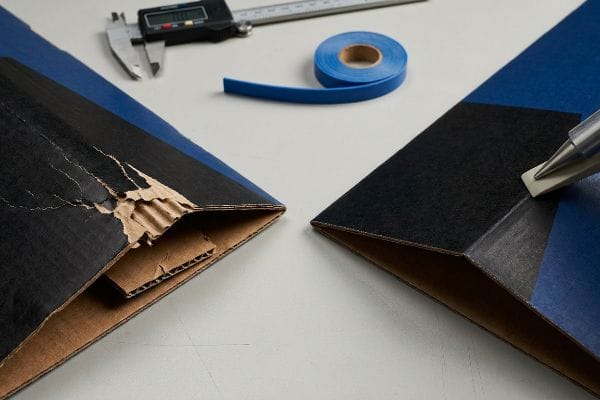 Damaged black corrugated board with litho-cracking exposing brown flutes next to a perfectly creased edge, with a caliper and blue tape.