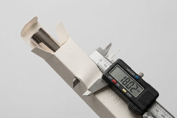 Caliper Measuring Cosmetic Carton A digital caliper measures an 18.02 mm cosmetic paperboard carton containing a cylindrical product, illustrating panel bulge for micro-clearance.