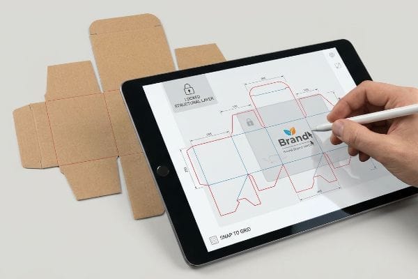 Packaging Dieline Design A hand uses a stylus on a tablet displaying a "Brandt" packaging dieline with "Locked Structural Layer" and "Snap to Grid", beside an unfolded cardboard box.