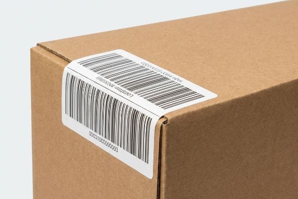 Brown corrugated box corner with a white logistics label, barcode, and 'Logistiken 70200003' text, creased around the fold.