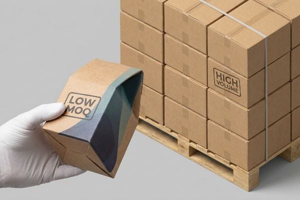 Low MOQ High Volume A gloved hand holds a custom Kraft box labeled "Low Moq", contrasting with a pallet of "High Volume" corrugated shipping boxes.