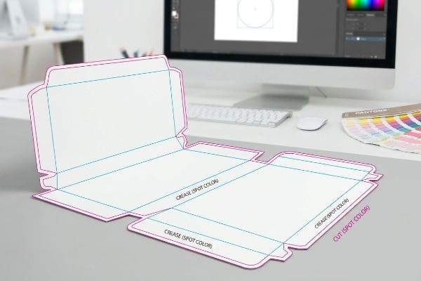 Spot Color Dieline White packaging dieline template showing magenta Cut (Spot Color) and cyan Crease (Spot Color) lines, on a desk with a monitor and Pantone guide.