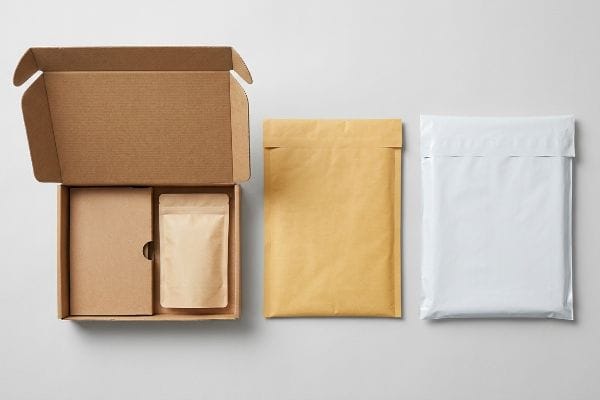 Various mailer package types, including an open corrugated box with internal packaging, a kraft padded envelope, and a white poly bag, arranged on a light surface.