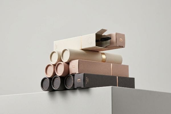 Hochwertige Kosmetikverpackungen in verschiedenen Ausführungen: cremefarbene, roségoldene und anthrazitfarbene Kartonschachteln und -tuben, eine davon geöffnet mit einem Mascara-Bürstchen.