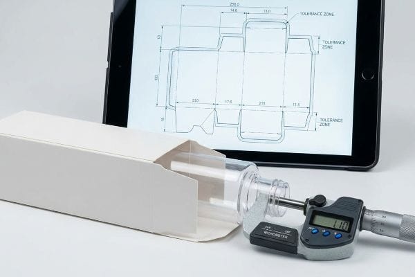 Packaging Dieline Micrometer Measurement Digital micrometer measures a clear cosmetic bottle, alongside an open white box and a tablet displaying a packaging dieline with Tolerance Zone details.