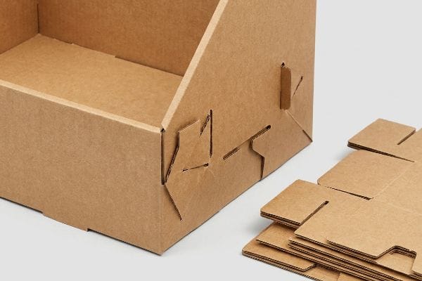 صندوق من الورق المقوى البني المموج مع ألسنة ورقية متشابكة ظاهرة لتجميع المواد الأحادية، بجانب كومة من قطع الكرتون المسطحة.