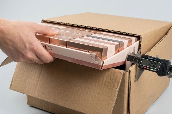 Corrugated cardboard shipping box with a hand inserting a Revlon Signature Eyeshadow retail tray, while a digital caliper measures the nested clearance.