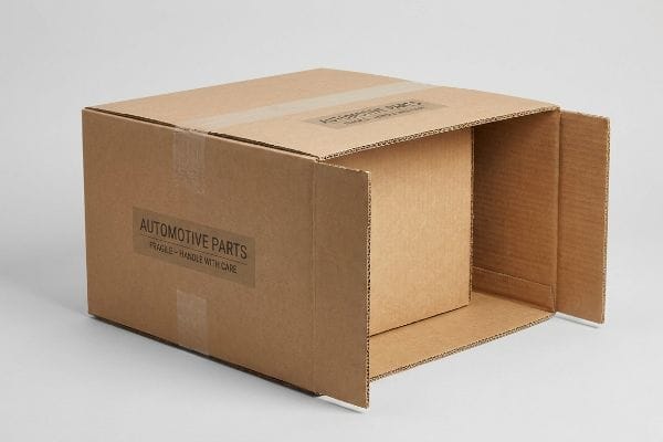 自動車部品輸送箱 茶色の段ボール製の輸送箱が部分的に開いており、内側の段ボール箱が見えている。箱には「自動車部品」と「壊れ物注意 - 取り扱い注意」とラベルが貼られている。.
