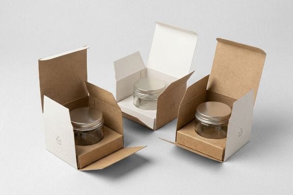 Drei offene Kosmetik-Displayboxen aus Kraftpapier, zwei mit heller Außenseite und brauner Innenseite, eine mit brauner Außenseite und weißer Innenseite, jede mit einem leeren Glasbehälter mit silbernem Deckel und einem dezenten „M“-Logo.