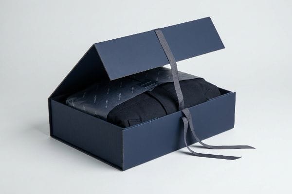 Premium Gift Box Packaging Navy blue premium rigid gift box, partially open, showcasing a folded garment in patterned tissue paper, secured by a grey ribbon.