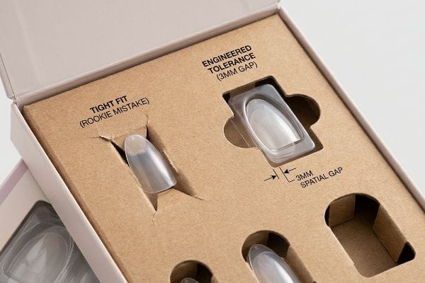 Packaging Tolerance Comparison Brown cardboard insert demonstrating cosmetic packaging engineering: "Tight Fit (Rookie Mistake)" versus "Engineered Tolerance (3mm Spatial Gap)" for press-on nails.