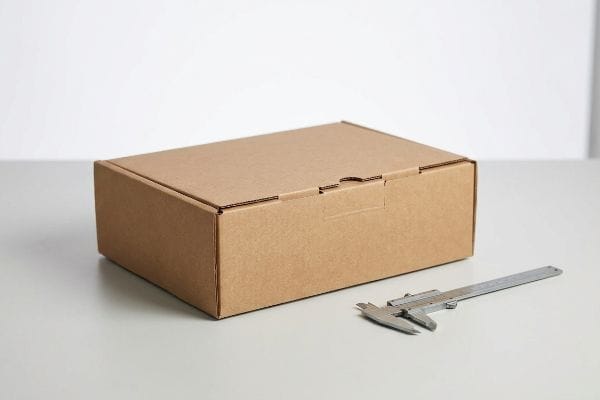 Packaging Box Caliper Measurement Brown corrugated box and silver caliper for precise packaging thickness measurement on a light gray surface.