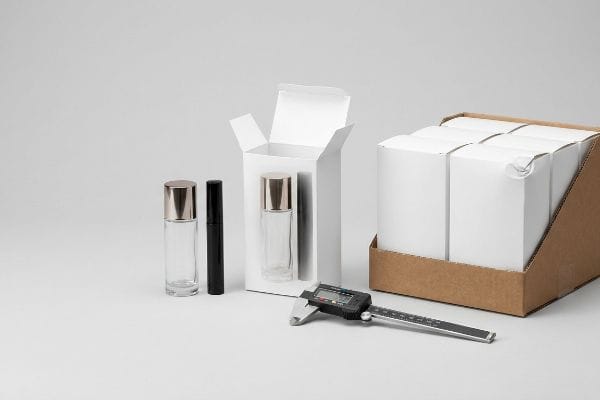 Clear glass foundation bottles, mascara tube, white folding cartons, digital caliper, and corrugated PDQ tray for cosmetics packaging.