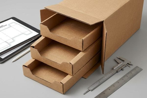 Corrugated Tray Design Raw corrugated cardboard box with three nested merchandising trays, a tablet showing a packaging CAD design, and a caliper for precise measurement.