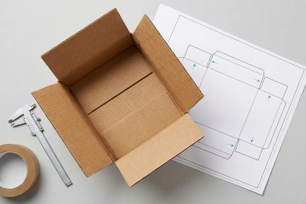 Custom Packaging Structural Design A workspace displays an open corrugated cardboard box, a caliper, tape, and a structural design blueprint for custom packaging.