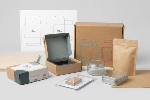 A collection of packaging samples featuring corrugated RSC and HSC dielines, a kraft box, flexible pouch, glass jar, metal tin, and a plastic blister pack.