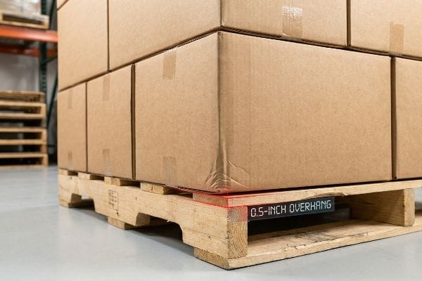 パレットの突き出し部分の測定 木製パレットの上に積み重ねられた段ボール箱。赤いレーザー線が0.5インチの突き出し部分を示しており、不適切な積載状態を示している。.
