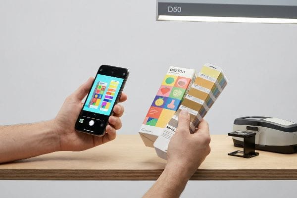 Pantone Color Verification Hands hold a smartphone showing digital color swatches and Pantone books under D50 light, with a spectrophotometer on a wooden table.