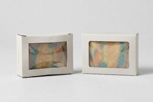 Two light beige paperboard boxes with clear PLA window films showcasing colorful marbled artisanal soap bars inside.