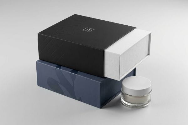 Black sleeve box with silver logo, geometric pattern, partially open white tray, on dark blue box, beside cosmetic jar.