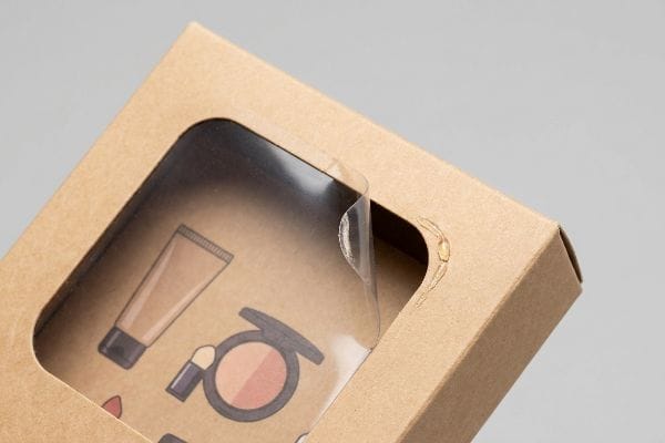 Peeling Window Patch Failure Brown kraft paperboard box with a die-cut window showing illustrated foundation and eyeshadow, a clear polymer film peeling from corner, highlighting adhesive failure.