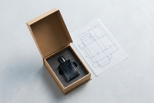 Custom Packaging Dieline An open kraft paper box holds a black perfume bottle in a foam insert, alongside a custom CAD dieline for packaging design.