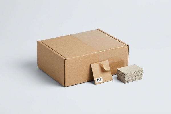 Brown cardboard box, a curled PLA laminated sample, and stacked paperboard squares, highlighting the PLA recycling trap.