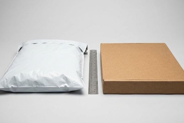 Dicke des Versandkartons Vergleich eines weißen Polybeutels und eines braunen Wellpappkartons mit einem Metalllineal zur Veranschaulichung der Verpackungsdicke für die Postklassifizierung.